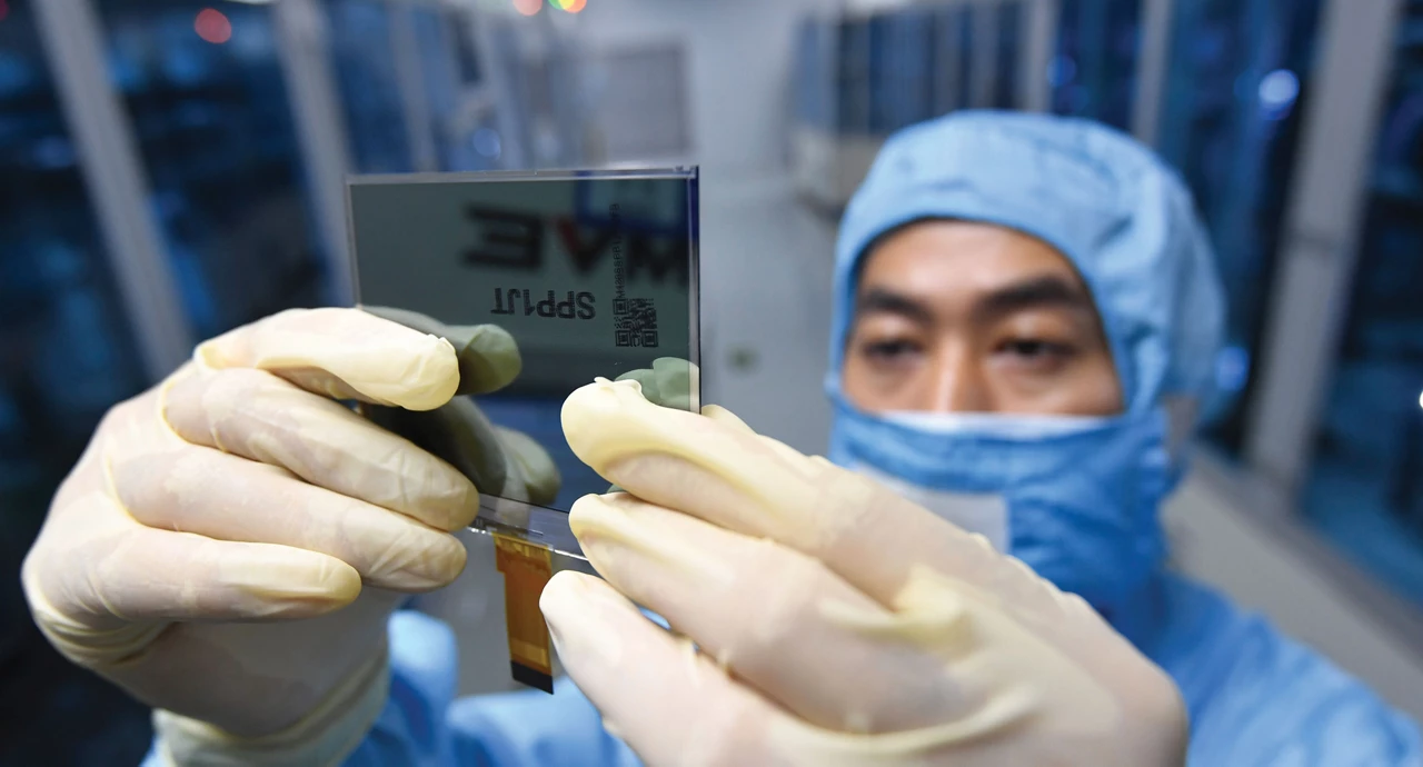A person wearing a blue clean room suit and white gloves holds up a square piece of glass.