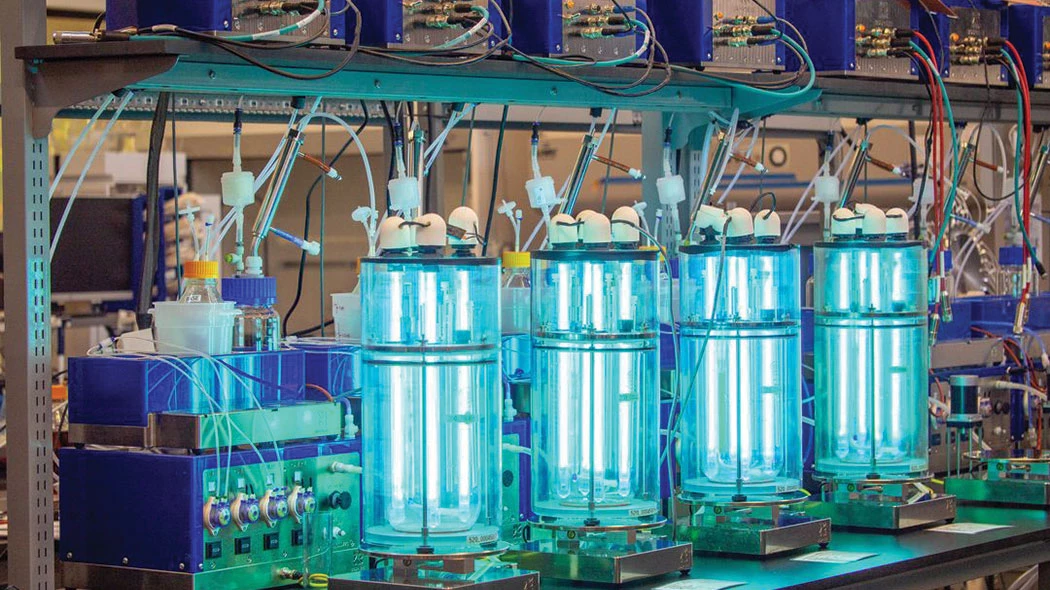 A line of glowing bioreactors sits on a lab bench surrounded by assorted wires and equipment.